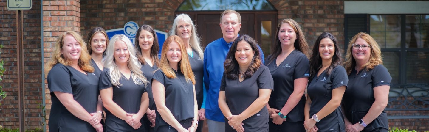Sutton Dentistry Group photo of a dental team standing outdoors in front of a brick building. The group includes a man in a blue shirt and women wearing black uniforms, all smiling at the camera.