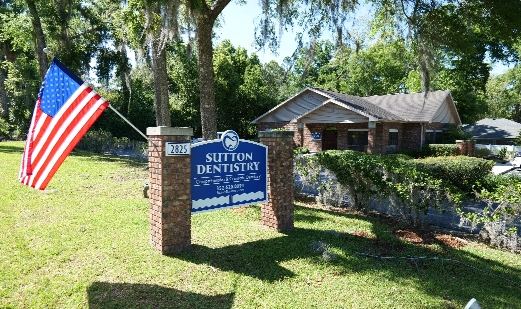 Sutton Dentistry Exterior view of Sutton Dentistry, featuring a brick sign with the clinic name and logo, an American flag on a pole, and a one-story building surrounded by trees.