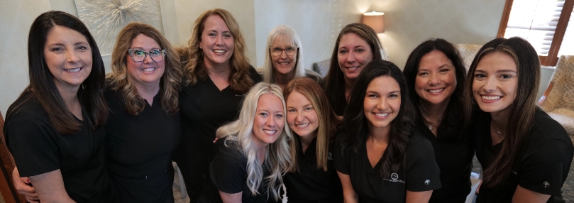 Sutton Dentistry Group photo of ten smiling female team members from Sutton Dentistry, wearing black uniforms, gathered in a warmly lit indoor space.