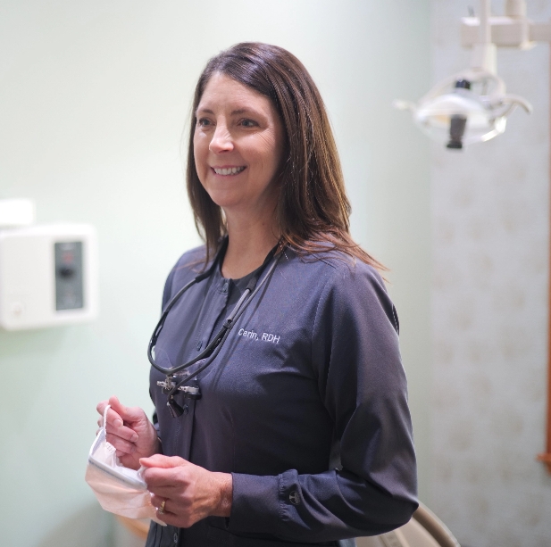 Sutton Dentistry Smiling dental hygienist in a dark gray uniform labeled 'Cami, RDH,' holding a mask in a dental treatment room with equipment in the background.